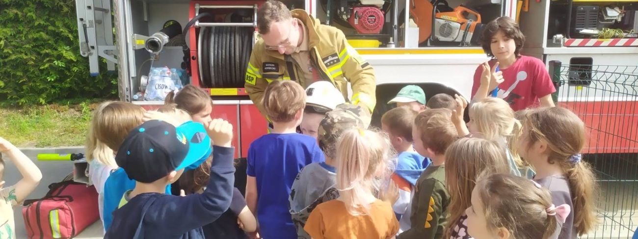 Strażak prezentuje grupie dzieci kask strażacki na tle wozu strażackiego