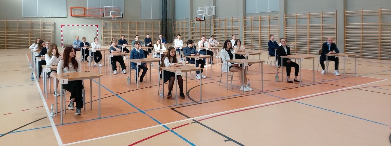 Uczniowie klasy ósmej siedzą przy stolikach na sali gimnastycznej