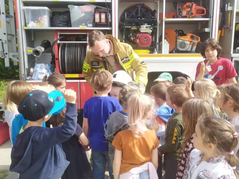Strażak prezentuje grupie dzieci kask strażacki na tle wozu strażackiego